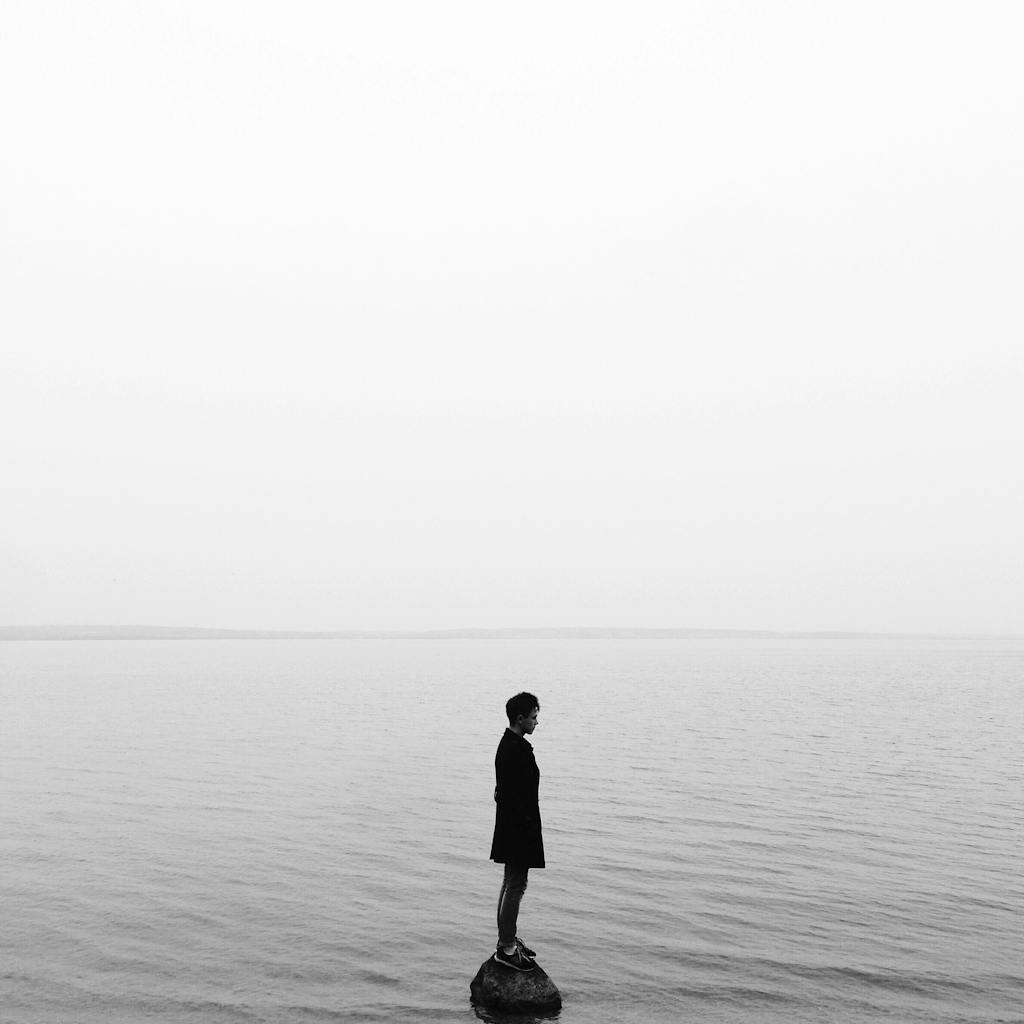 Black and white image of a solitary person on a rock in a calm lake, evoking solitude and reflection.